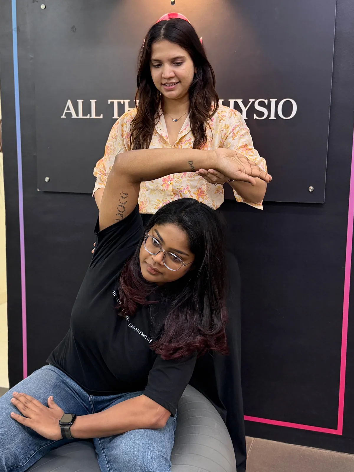 Assisted shoulder stretch in front of the All Things Physio logo wall